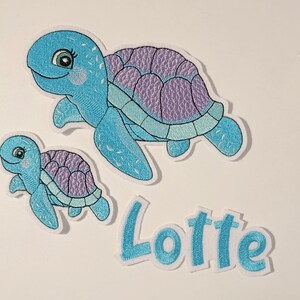 Könnte beinhalten: Zwei aufgestickte Aufnäher mit Meeresschildkröten in Blau- und Lilatönen. Eine größere Schildkröte befindet sich über einer kleineren, darunter der Name "Lotte" in Blau. Die Aufnäher haben einen weißen Rand.
