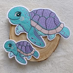 Könnte beinhalten: Zwei gestickte Schildkrötenaufnäher. Die größere Schildkröte ist türkis und lila mit einem rosa Bauch und weißem Rand. Die kleinere Schildkröte hat das gleiche Design. Beide liegen auf einer Holzscheibe.