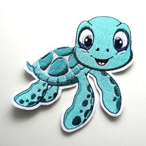 Könnte beinhalten: Ein gestickter Aufnäher mit einer Cartoon-Meeresschildkröte mit hellblauer Haut, einem dunkelblauen Panzer mit weißen Akzenten und großen schwarzen Augen. Die Schildkröte hat einen lächelnden Mund mit einer kleinen roten Zunge und dunklen Flecken.