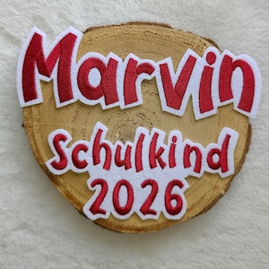 Könnte beinhalten: Eine Holzscheibe mit dem Namen "Marvin" in rot-weiß gestickten Buchstaben. Darunter sind die Worte "Schulkind 2026" ebenfalls rot-weiß gestickt. Das Holz hat eine natürliche, hellbraune Farbe.
