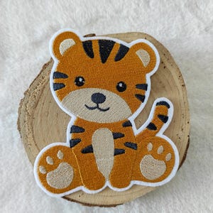 Könnte beinhalten: Ein niedlicher, orangefarbener und schwarzer gestickter Tiger-Applik mit weißen Nähten. Der Tiger sitzt mit sichtbaren Pfoten und hat einen freundlichen Ausdruck.