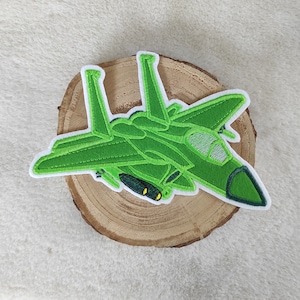 Könnte beinhalten: Ein grüner, gestickter Aufnäher in Form eines Kampfflugzeugs. Der Aufnäher hat einen weißen Rand, detaillierte Nähte und ein dunkelgrünes Cockpit. Das Flugzeug wird auf einer Holzscheibe präsentiert.