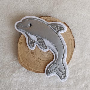 Könnte beinhalten: Ein grauer Delfin-Aufnäher mit weißem Rand, auf einer Holzscheibe. Der Delfin ist detailliert gestickt und zeigt eine geschwungene Form und ein lächelndes Gesicht. Geeignet für Kleidung oder Accessoires.