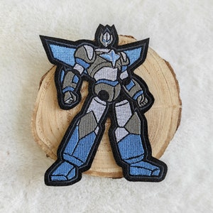 Könnte beinhalten: Gestickter Aufnäher mit einem stilisierten Roboter in Blau-, Grau- und Schwarztönen. Der Roboter hat ein geometrisches Design mit schwarzer Umrandung. Der Aufnäher liegt auf einer Holzscheibe vor weißem Hintergrund.