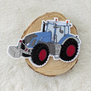 Könnte beinhalten: Blau-grauer gestickter Traktor-Patch mit roten Rädern. Der Traktor hat einen Frontlader und befindet sich auf einem hölzernen Hintergrund.