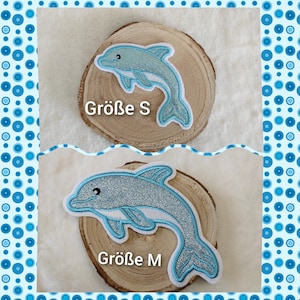 Könnte beinhalten: Zwei blaue Glitzer-Delfin-Applikationen auf Holzrundlingen. Der kleinere Delfin ist mit "Größe S" und der größere Delfin mit "Größe M" beschriftet.