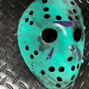 May include: A teal hockey mask with black accents and purple markings. The mask has numerous circular eye and mouth holes. The mask is on a black diamond plate surface.