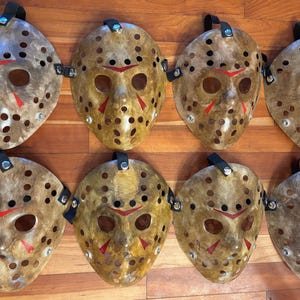 May include: Eight hockey-style masks with a weathered, distressed appearance. The masks are primarily tan with red accents around the mouth and eyes. Each mask has a black strap and is arranged on a wooden surface.