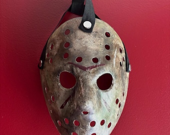 May include: A weathered hockey mask with a black strap and rivet detail. The mask has numerous circular eye and mouth holes, with a distressed, aged appearance. Red paint accents the forehead and mouth area. The mask is suspended against a red background.