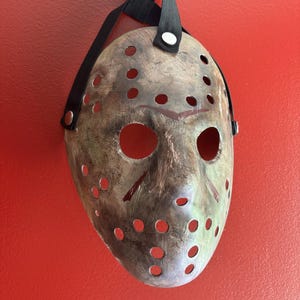 May include: A weathered hockey mask with numerous circular holes and large eye openings. The mask has a distressed, metallic appearance with red accents and is suspended by a black strap. The mask is against a red background.
