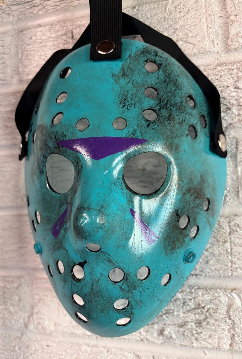 May include: A teal hockey mask with a black strap and eye holes. The mask has a purple triangle above the eyes and purple markings on the cheeks. The mask has a distressed look with black smudges.