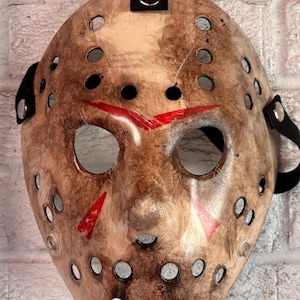 May include: A weathered hockey mask with a distressed finish, featuring red markings and numerous circular eye and ventilation holes. The mask has black straps for securing it to the face, and is a prop or costume piece.