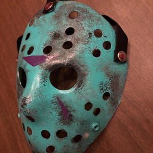 May include: A teal hockey mask with black and purple accents. The mask has numerous circular eye and mouth holes, and a black strap with metal rivets. The mask is designed with a distressed, weathered appearance.