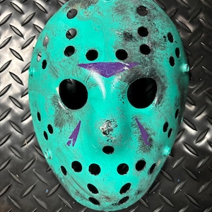 May include: A teal hockey mask with black accents and purple markings. The mask has numerous black circular eye and ventilation holes. The mask is a prop or costume piece.