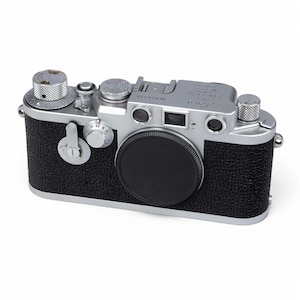Könnte beinhalten: Eine silber-schwarze Leica Messsucherkamera mit einem strukturierten schwarzen Kunstledergehäuse, einem runden Objektivdeckel, mehreren metallischen Einstellrädern auf der Oberseite, einem Sucherfenster und Gravuren, einschließlich des Markennamens Leica.