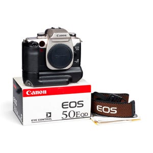 以下が含まれることがあります： シルバーとブラックのCanon EOS 50Eカメラボディとバッテリーグリップ。EOS 50EQDと記された白と赤の元箱の上に置かれています。EOSの白い文字が入った茶色と黒の織り込みカメラストラップが、紙の取扱説明書と共に箱の横に配置されています。