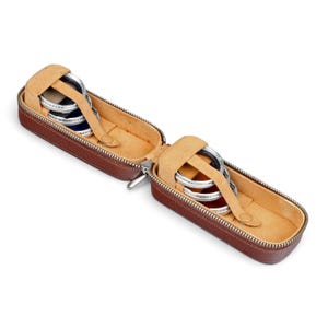 May include: A brown leather-textured zipped case with a tan interior, containing two sets of circular metal camera filters. Each set is secured by a tan leather strap with a snap button. The silver-rimmed filters feature text markings.