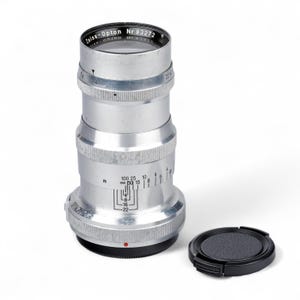May include: A silver Zeiss-Opton camera lens with a black lens cap. The lens has various measurement markings and the text "Zeiss-Opton Nr 93272" printed on the top. The lens cap is round and textured.