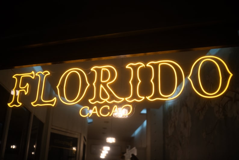 Puede incluir: Un letrero de ne&oacute;n amarillo brillante con la palabra "FLORIDO" en letras grandes y "CACAO" debajo. El letrero est&aacute; sobre una entrada de tienda, con reflejos en el cristal.