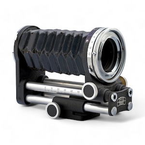 Puede incluir: Un objetivo de cámara de fuelle negro con un anillo de enfoque plateado y una base plateada y negra con un riel deslizante y una escala marcada en centímetros. La base está etiquetada como "Zeiss Ikon".