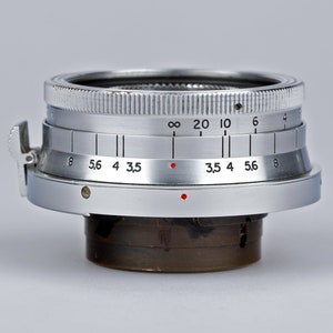 Puede incluir: Primer plano de una lente de c&aacute;mara plateada con marcas en blanco y negro y puntos rojos. La lente tiene un borde estriado y un dial con n&uacute;meros. Lente para fotograf&iacute;a.