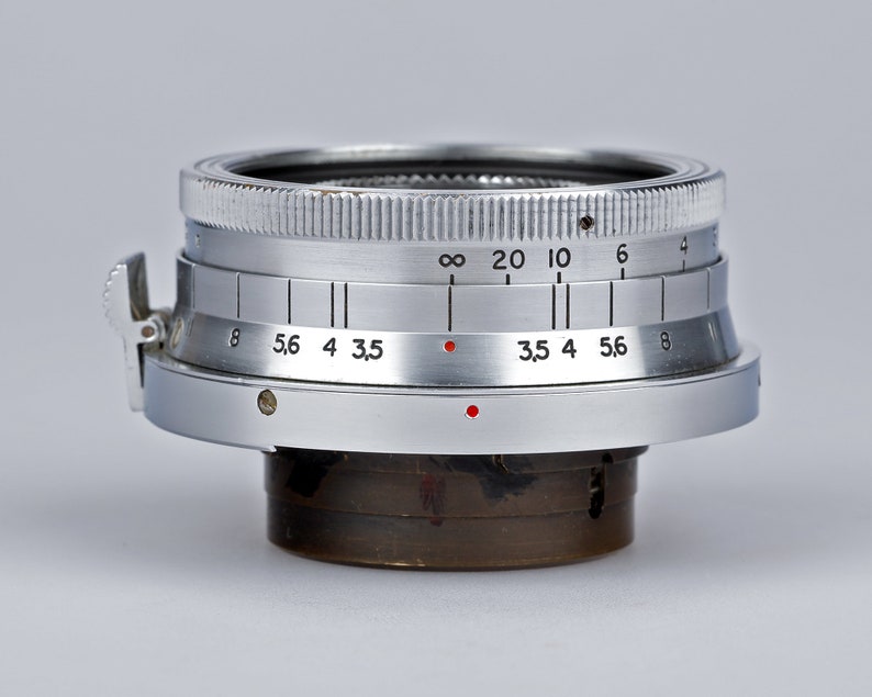 Puede incluir: Primer plano de una lente de c&aacute;mara plateada con marcas en blanco y negro y puntos rojos. La lente tiene un borde estriado y un dial con n&uacute;meros. Lente para fotograf&iacute;a.