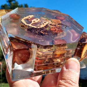 May include: Octagonal clear resin coaster with embedded wood and metal pieces. The coaster has a layered appearance with different shades of brown and gold.