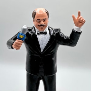 May include: A black and white action figure of a man wearing a black tuxedo with a white shirt and a black bow tie. He is holding a silver microphone with a blue and yellow "WF" logo. He is smiling and has a mustache.