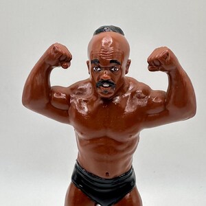 Pode incluir: Uma estatueta marrom de um homem musculoso flexionando os braços. A figura está usando cuecas e sapatos pretos. A estatueta tem uma cabeça careca com um pequeno tufo de cabelo preto e um bigode.