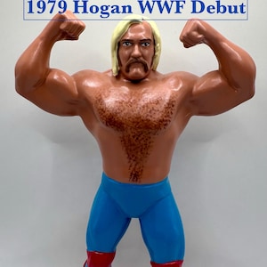 May include: A vintage action figure of a muscular wrestler in a classic pose. The figure has blonde hair, a mustache, and is wearing blue tights and red boots. The text "1979 Hogan WWF Debut" is displayed at the top.
