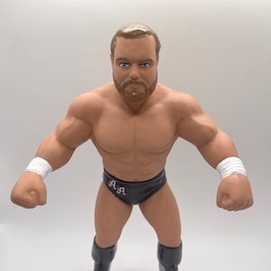 May include: A wrestling action figure with a brown beard, blue eyes, and a black wrestling outfit with the letters "A.A." on the front. The figure is posed with fists clenched and is wearing black boots.
