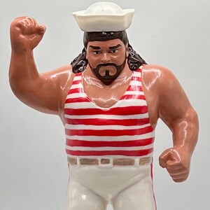 Pode incluir: Uma figura de ação de lutador vestindo um chapéu de marinheiro branco, uma camiseta regata listrada de vermelho e branco e calças brancas com listras vermelhas nas laterais.