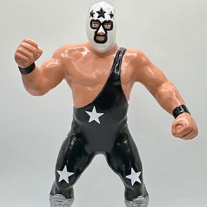 May include: A vintage action figure of a wrestler in a black singlet adorned with white stars. The figure has a white mask with black accents and silver boots. The wrestler is posed with a clenched fist.