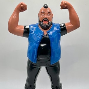 May include: A blue and black action figure of a wrestler with a mohawk hairstyle. The figure is wearing a blue vest and black pants with black boots.