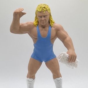 May include: A blue wrestling figure with blonde hair, wearing white boots and holding a white towel.