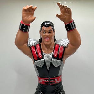 May include: A figurine of a muscular wrestler in a black and red outfit with silver accents. The figure has black boots, red knee pads, and wristbands. The wrestler is posed with arms raised.