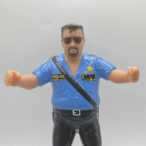 May include: A figurine of a man in a blue police uniform with the text "THE BOSS MAN" on the shoulder. The figure is wearing sunglasses and black pants. A black baton lies at the figure's feet.
