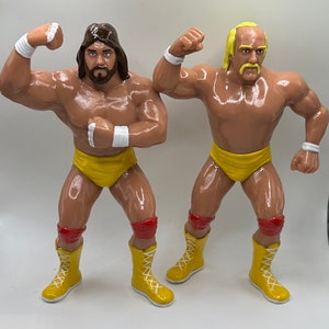 May include: Two wrestling action figures, one with brown hair and a yellow and red outfit, the other with blonde hair and a yellow and red outfit.