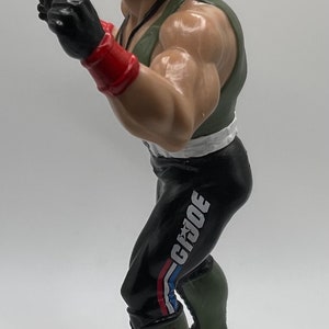 May include: A green and black action figure of a soldier wearing a green hat with a red and white band. The figure is wearing black pants with a white and red stripe and the word "G.I.JOE" printed on the side. The figure is wearing green boots with a gold star on the side.
