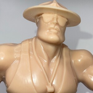 May include: A tan plastic action figure wearing a tan hat and sunglasses. The figure has a necklace with a whistle attached.