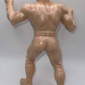 May include: A tan plastic action figure of a man wearing a tan hat and a tan vest. The figure is posed with one arm raised and the other arm bent at the elbow. The figure is standing on a white surface.