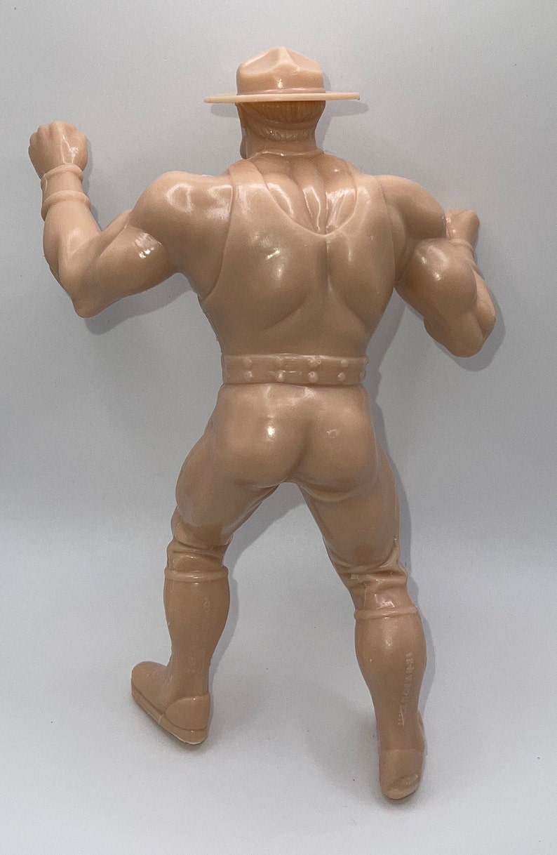 May include: A tan plastic action figure of a man wearing a tan hat and a tan vest. The figure is posed with one arm raised and the other arm bent at the elbow. The figure is standing on a white surface.