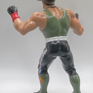 May include: A green and black action figure wearing a green tank top, black pants with a white stripe and a red, white, and blue stripe, and a green and black hat. The figure is holding its fists up in a fighting pose.