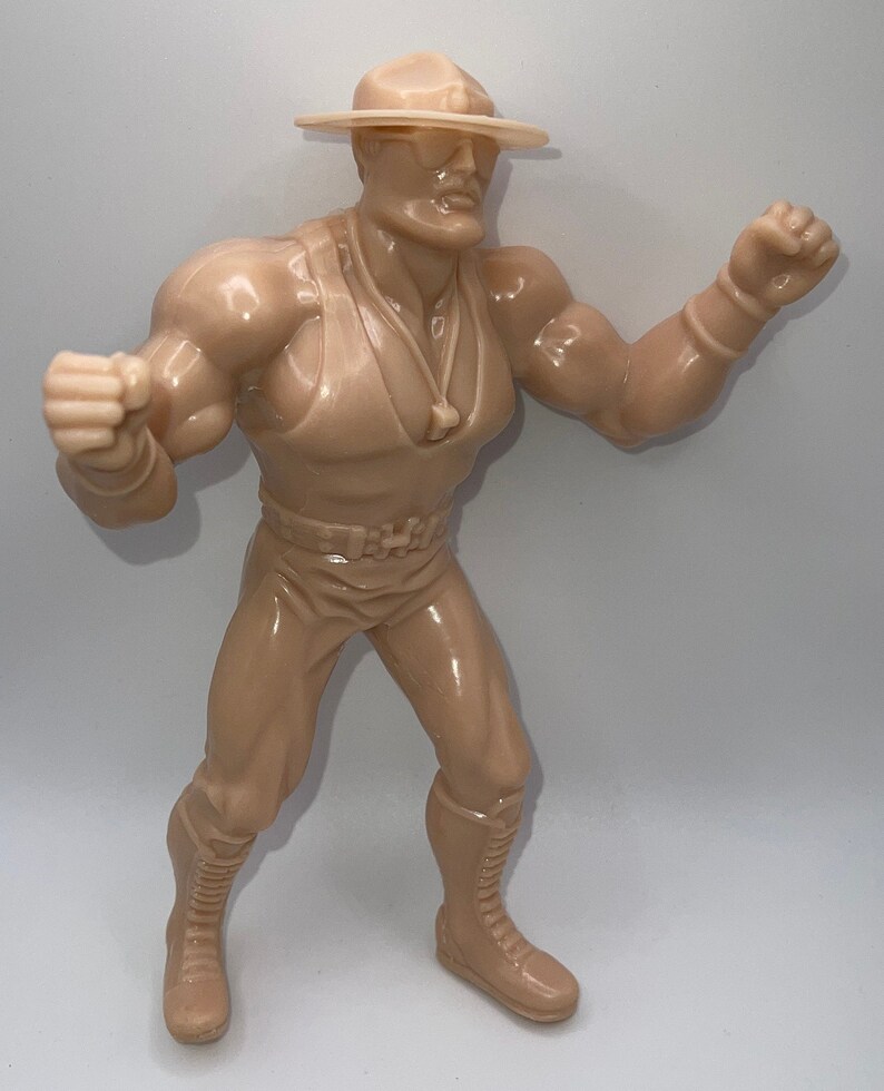 May include: A tan plastic action figure of a wrestler wearing a brown hat, sunglasses, a brown vest, and brown pants. The figure is posed with one arm raised and one fist clenched.