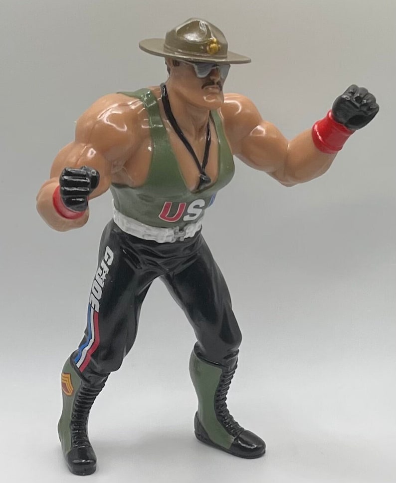 May include: A green and black action figure wearing a green and brown military hat with a gold star. The figure is wearing a green tank top with the letters "US" on the front. The figure is also wearing black pants with the word "G.I.JOE" on the side. The figure is holding its fists up in a fighting stance.