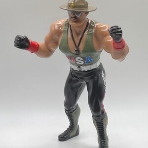 May include: A green, black, and red action figure of a wrestler wearing a military-style hat, sunglasses, and a tank top with the word "USA" on it. The figure is holding its fists up in a fighting pose.