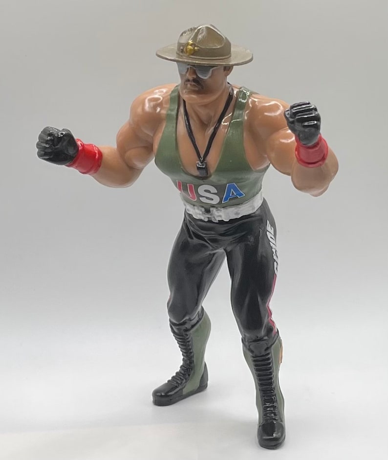 May include: A green, black, and red action figure of a wrestler wearing a military-style hat, sunglasses, and a tank top with the word "USA" on it. The figure is holding its fists up in a fighting pose.