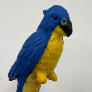 May include: A blue and yellow plastic toy parrot with a black beak. The parrot is perched on a branch.