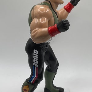 May include: A green and black action figure with a green hat and red and black gloves. The figure is wearing black pants with a white "G.I. JOE" logo on the side. The figure is posed in a fighting stance.