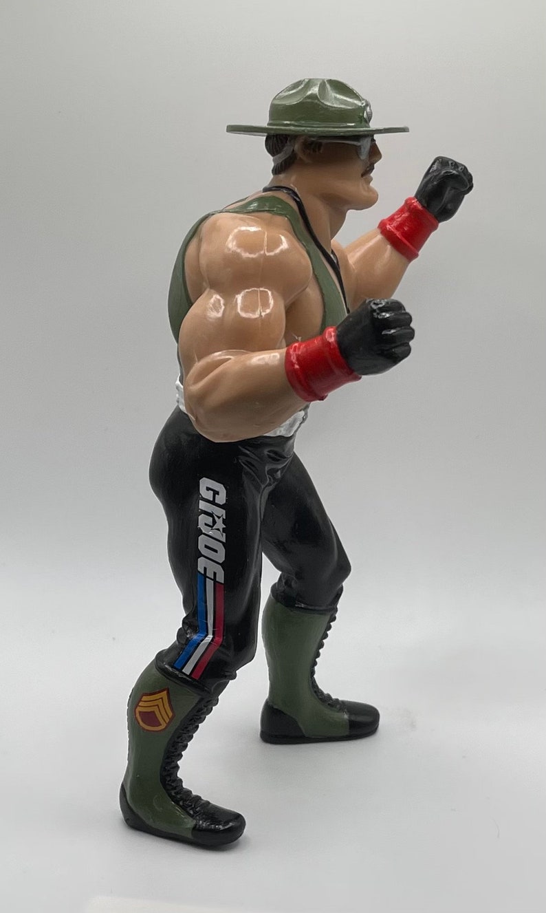 May include: A green and black action figure with a green hat and red and black gloves. The figure is wearing black pants with a white "G.I. JOE" logo on the side. The figure is posed in a fighting stance.